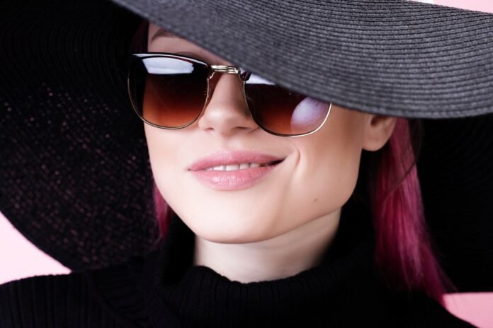 A young woman with a small face wearing stylish tortoiseshell sunglasses, standing outdoors in natural light, with a scenic green background, showcasing a flattering sunglass style for petite facial features.